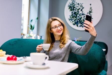 Young pretty woman at indoor cafe is drinking coffee with dessert, smiling and making selfie. Travel concept.の写真素材