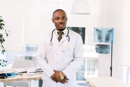 Young handsome friendly african american man doctor in white coat standing in modern medical office. Light modern clinic, radiography on the backgroundの写真素材