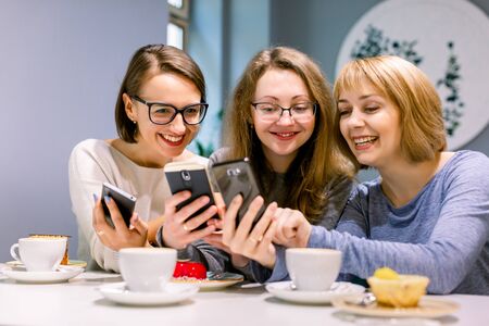 Group of three young women smiling and showing the pictures on the smartphones each other, sitting in restaurant, drinking coffee and eating dessertsの写真素材