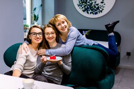 Women friends sitting on the sofa in cafe, looking at camera, one girl holds plate with delicious red dessertの写真素材