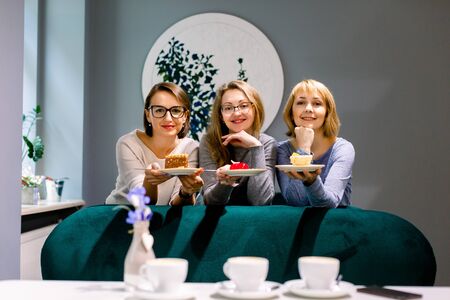 Three pretty women friends holding plates with colorful cakes looking at camera in cafe indoors. Picture presenting group of friends chatting in cafeの写真素材