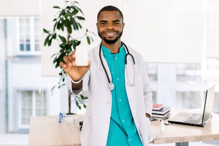 Young African Male Doctor with Stethoscope and in medical coat Holding medicines for treatment patients, smiling and posing in modern clinic. Focus on faceの写真素材
