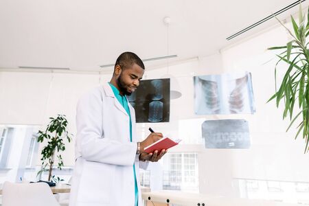 Medicine, oncology, tumour. Professional African male oncologist looking at the X-ray photos of the patients and making notes in red notebookの写真素材