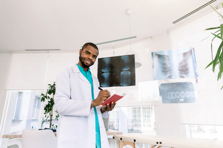 health and medicine concept - smiling African man doctor studying x-ray of the patients while standing in modern medical officeの写真素材