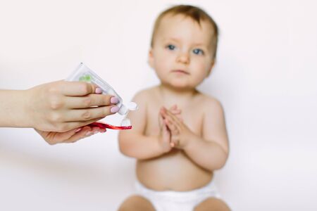 Cute little baby girl waits for the brushing teeth with red toothbrush. Isolated on white background. Mother holds toothbrush and toothpaste. Dental hygieneの写真素材