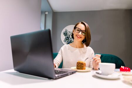 Concentrated at work. Confident smiling young woman in smart casual wear working on laptop while sitting near window in creative office or cafe, eating dessert and drinking coffeeの写真素材