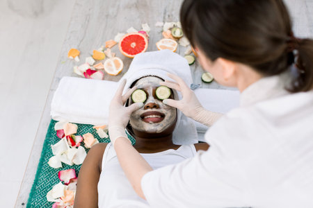 Female cosmetologist in uniform and protective gloves doing cucumber mask on eyes for relaxation in beauty salon. Smiling African woman in white towel lying on carpet and enjoying procedureの写真素材