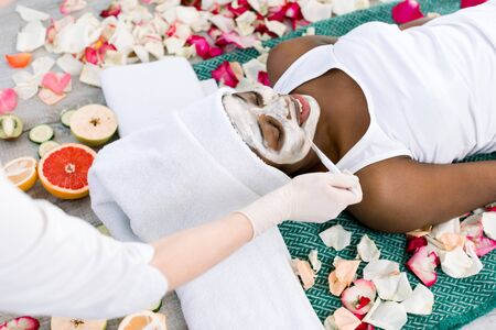 Beautiful african girl resting relaxing in spa resort with closed eyes while cosmetologist applies facial mask on her faceの写真素材