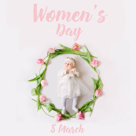 A little beautiful newborn girl in a white dress is lying on the bed among the flowers and the inscription Women's Day. 8 March. Greeting cardの写真素材