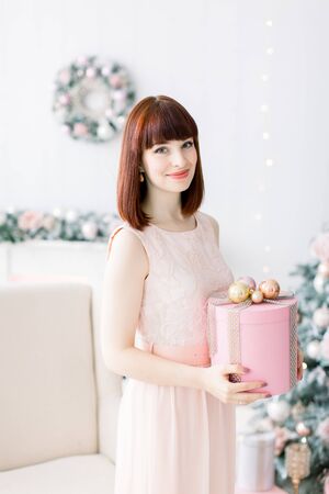 Smiling pretty young woman in light orange pink dress holding christmas present box and standing in beutiful cozy living room with Christmas decorationsの写真素材