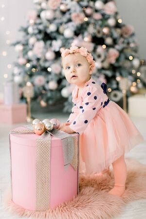 Cute little baby girl in pink dress standing near the beautiful Christmas box on the background of Christmas tree and decorations. Christmas, family, New Year conceptの写真素材