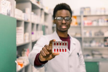Portrait of positive African American man pharmacist in modern drugstore holding red pills in the hand. Focus on the hand.の写真素材