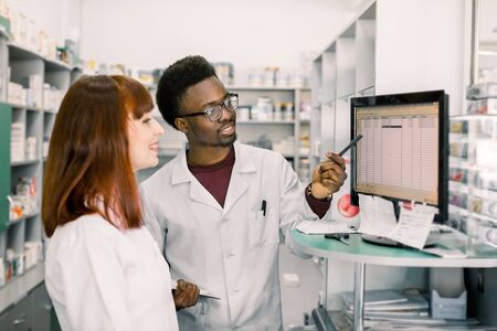 Portrait of a handsome African pharmacist and his female colleague, at the counter of a drugstore, working together on computer.の写真素材