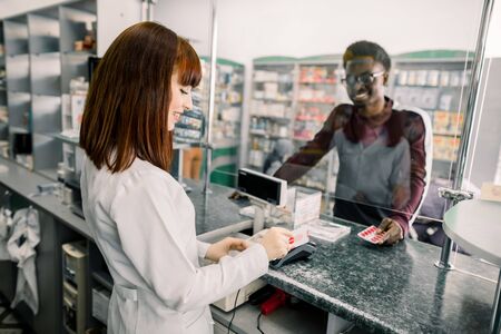 Young African man paying for pills with credit card in pharmacy. Smiling woman pharmacist giving pills to black man in drugstore.の写真素材
