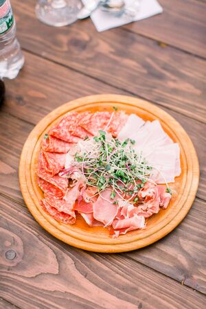 Food wooden plate with delicious salami, pieces of sliced ham, sausage, and greenery. Meat platter with selection. Cutting sausage and cured meat on a celebratory table.の写真素材