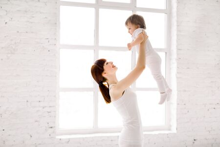 happy charming sporty mother in white wear holding cute little baby girl up overhead, while standing against big window in light hall gymの写真素材