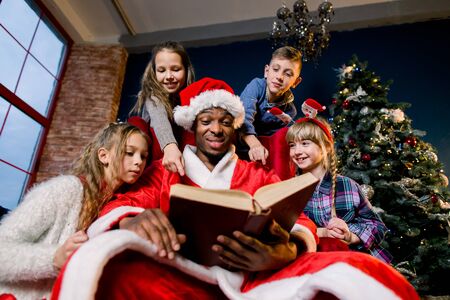 Small beautiful children listen carefully to the story Santa Claus reads while sitting on a red chair on the background of a Christmas tree.の写真素材
