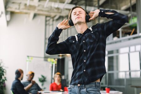 Happy young Caucasian businessman in casual wear with earphones listening music and having break in office. His succesful team working and discussing on the backgroundの写真素材
