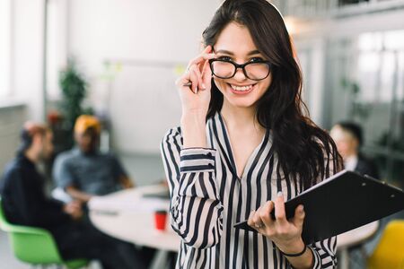 Young attractive designer woman in glasses smiling at camera in creative office while her colleagues sitting at the table and talking on the backgroundの写真素材