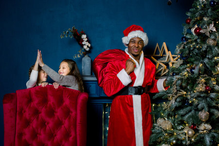 African Santa Claus puts gifts under a Christmas tree, and does not see that two little girls do not...