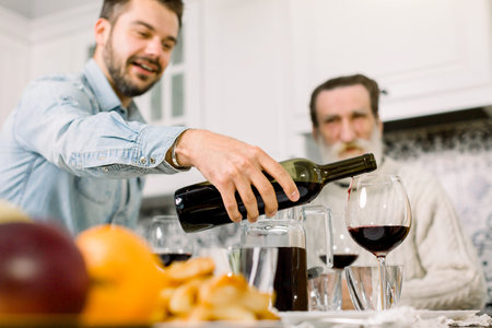 Closeup of young man holding bottle of red wine and pouring in glasses, festive table, traditional...