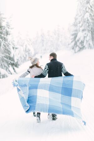 Young couple in love having a walk in winter snowy forest and holding warm wool blanket in handsの写真素材