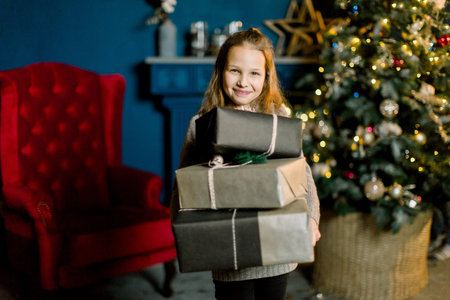 Little happy beautiful girl holds christmas gifts standing on the background of decorated Christmas...