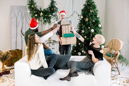 Caucasian young men presenting christmas gifts to friends while celebrating Christmas or New Year together. African boy and pretty Caucasian girls sitting on the sofa and drinkink champagne.の写真素材