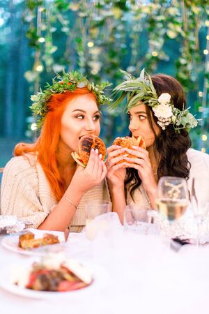 attractive funny and hungry young bride and her bridesmaid in green floral wreath on heads eating hamburgers, while sitting in stylish rustic restaurant in forest outdoorsの写真素材