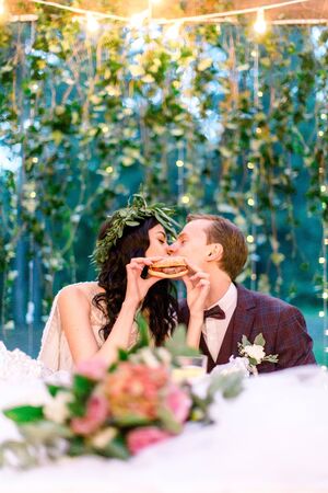 Stylish wedding couple eating fastfood and kissing, bride holds tasty burger in hands. Wedding outdoors.の写真素材