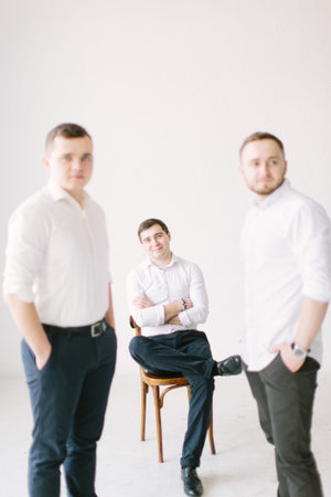 Stylish handsome groom sitiing on the chair posing while groomsmen standing near him, helping him to get ready in the morning. wedding preparation. black and white photoの写真素材