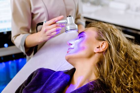Skin Cosmetology. Woman beautician Doing Blue Light Therapy On face of pretty young blond woman.の写真素材