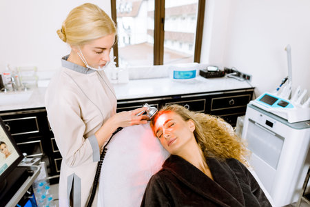 Female doctor conducts procedure for rejuvenating facial skin. Beautiful blond woman receiving facial treatment, removing pigmentation at clinic. Cosmetology light equipment. Anti age and wrinkle.の写真素材