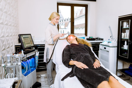 Woman doctor doing red led light therapy to young woman in beauty clinic, facial photo therapy for skin. Anti-aging treatments, photo rejuvenation procedureの写真素材