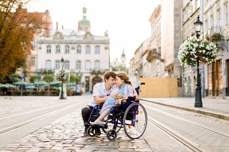 Couple in love in old city center. Pretty young girl with disease on a wheelchair and her lovely man, walking, smiling and touching foreheads, closed eyes.の写真素材