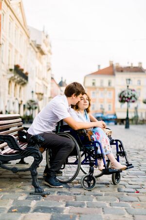 Young man hugs his pretty woman on the wheelchair from behind, while sitting on the bench on the street of old city.の写真素材