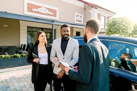 Young Caucasian man car dealer explaining sales contract to couple in business wear, african man and caucasian woman, buying a car, standing outdoors in auto salonの写真素材