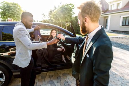 Caucasian young salesman handling keys to young African businessman after buying a car. Young Caucasian business lady is sitting in the carの写真素材
