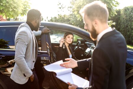 Happy multi ethnic couple, African man and Caucasian woman, buying the car, black crossover, woman is sitting in the car and holding car keys. Young salesman holds folder with contract for saleの写真素材