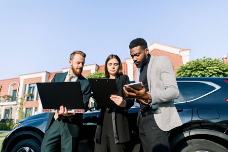 Young confidence business partners, African man and Caucasian man and woman, are working using tablet and laptop together before business trip standing near the black car outdoors.の写真素材