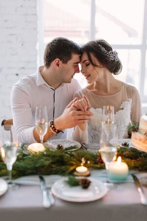 Bride and groom posing at the decorated banquet table in the white hall. Hand in hands. Enjoy a moment of happiness and love. A series of photos in my portfolio. Wedding flowers on a tableの写真素材