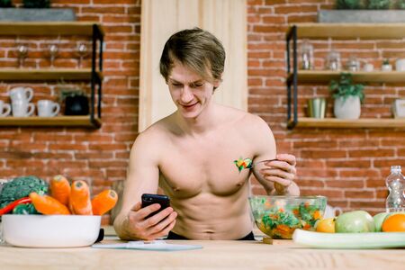 Close-up portrait of a handsome man eating salad with fresh vegetables and using smartphone on the red brick wall kitchen background. Healthy and vegan food concept.の写真素材