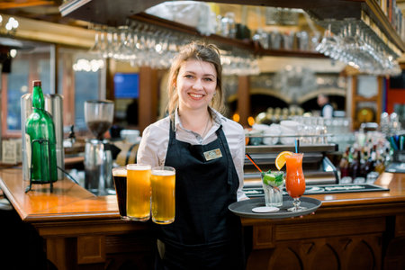 Smiling friendly waitress serving a pint of draft beer in a pub. Portrait of happy young woman serving beer in bar, looking at camera smilingの写真素材