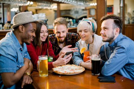 Group of young cheerful friends are sitting in a cafe, eating, drinking drinks. Friends take selfies...