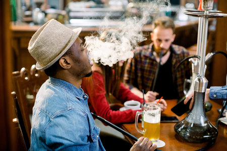 Cheerful company of friends, drinking beer and smoking hookah in shisha lounge. Rest in hookah,...