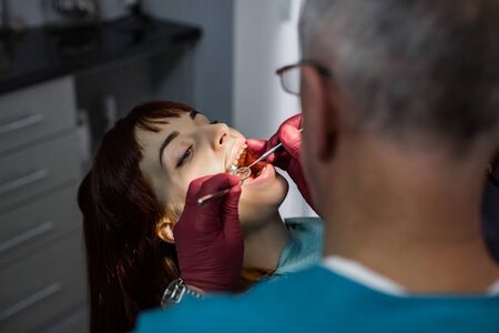 Side view of senior male doctor dentist at dentist surgery having tooth operation for young woman patient in modern stomatology clinic. Focus on woman faceの写真素材