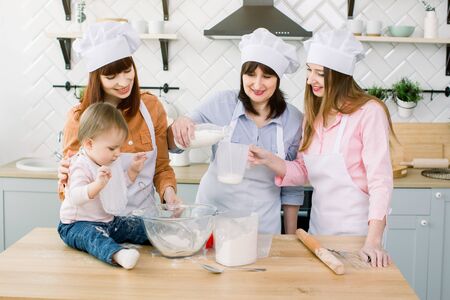 Happy family baking in the kitchen. Grandmother with her daughters and granddaughter preparing the dough, Granny pours milk from a bottle into a measuring glassの写真素材