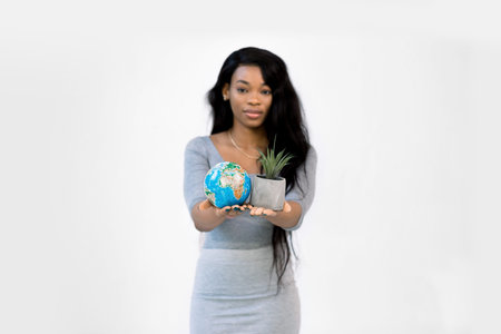 Young African American woman showing little earth globe in one hand and little gray pot with plant in another, isolated on white background. Focus on handsの写真素材