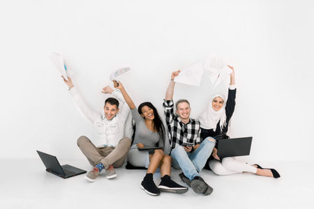 Excited group of four multiethnic friends keepeing hands raised, sitting together on the floor on...