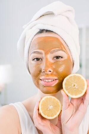 Beauty Skin Care Concept. Pretty Caucasian woman in white towel with brown mud facial mask on face holds citrus fruit on her hand on light background. Spa procedures and mask on skin.の写真素材